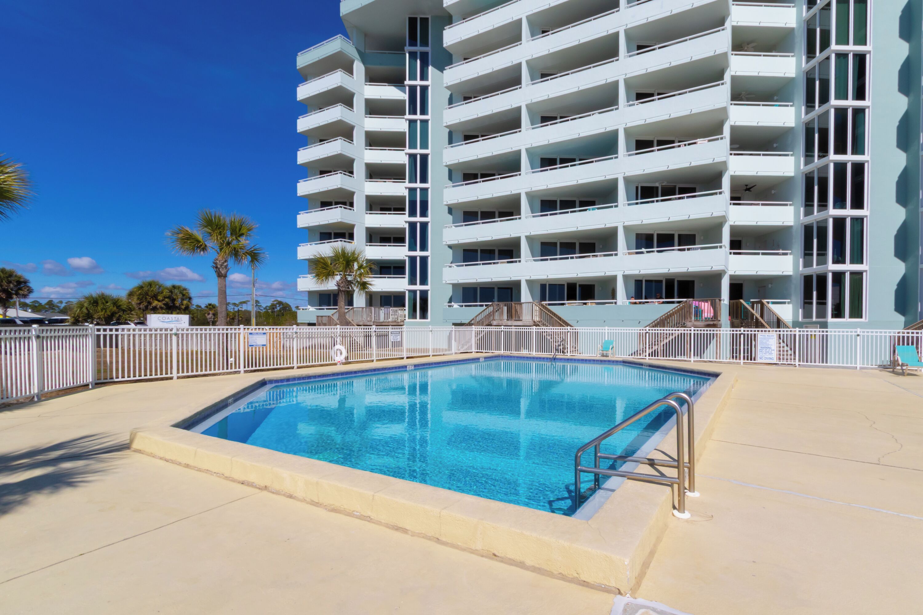 Une piscine entourée d’une clôture blanche, un immeuble à plusieurs étages doté de balcons, et des palmiers en arrière-plan.