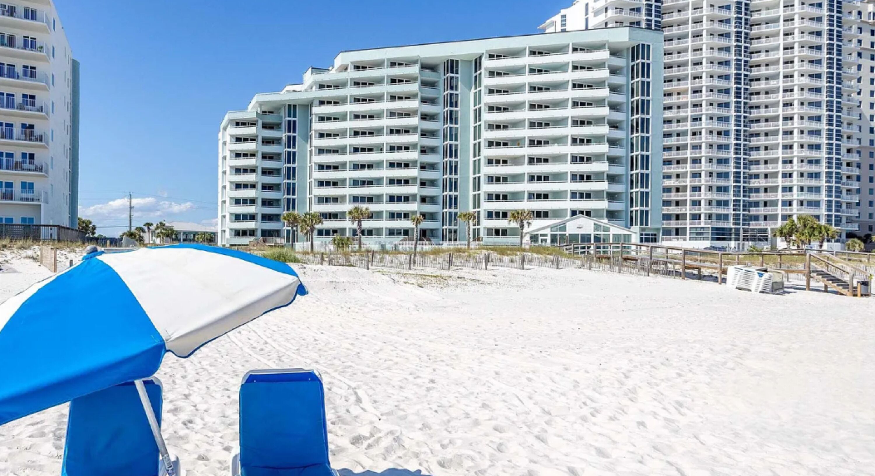 Ein Strandhotel mit mehreren Hochhäusern, einem Sandstrand und einem blau-weißen Sonnenschirm.