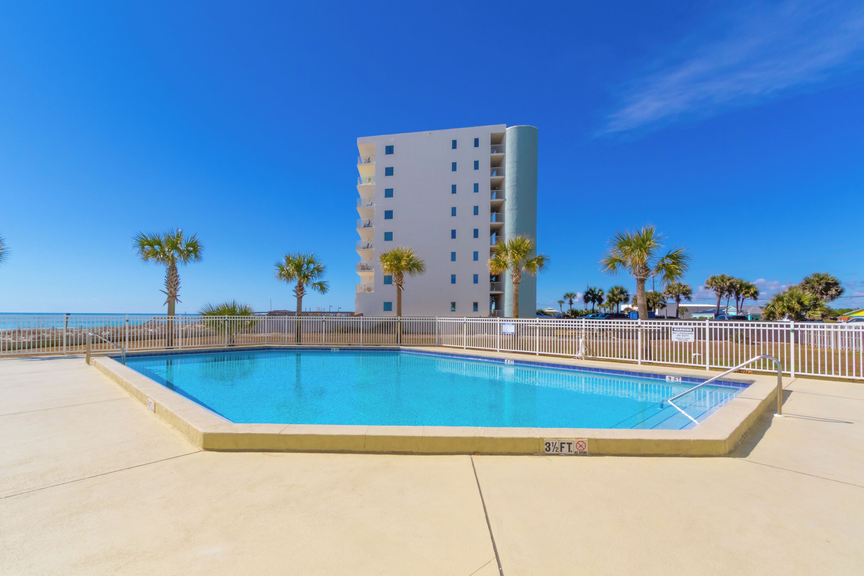 A rectangular swimming pool with a 3½ ft depth sign, surrounded by a white fence, palm trees, and a tall building in the background.
