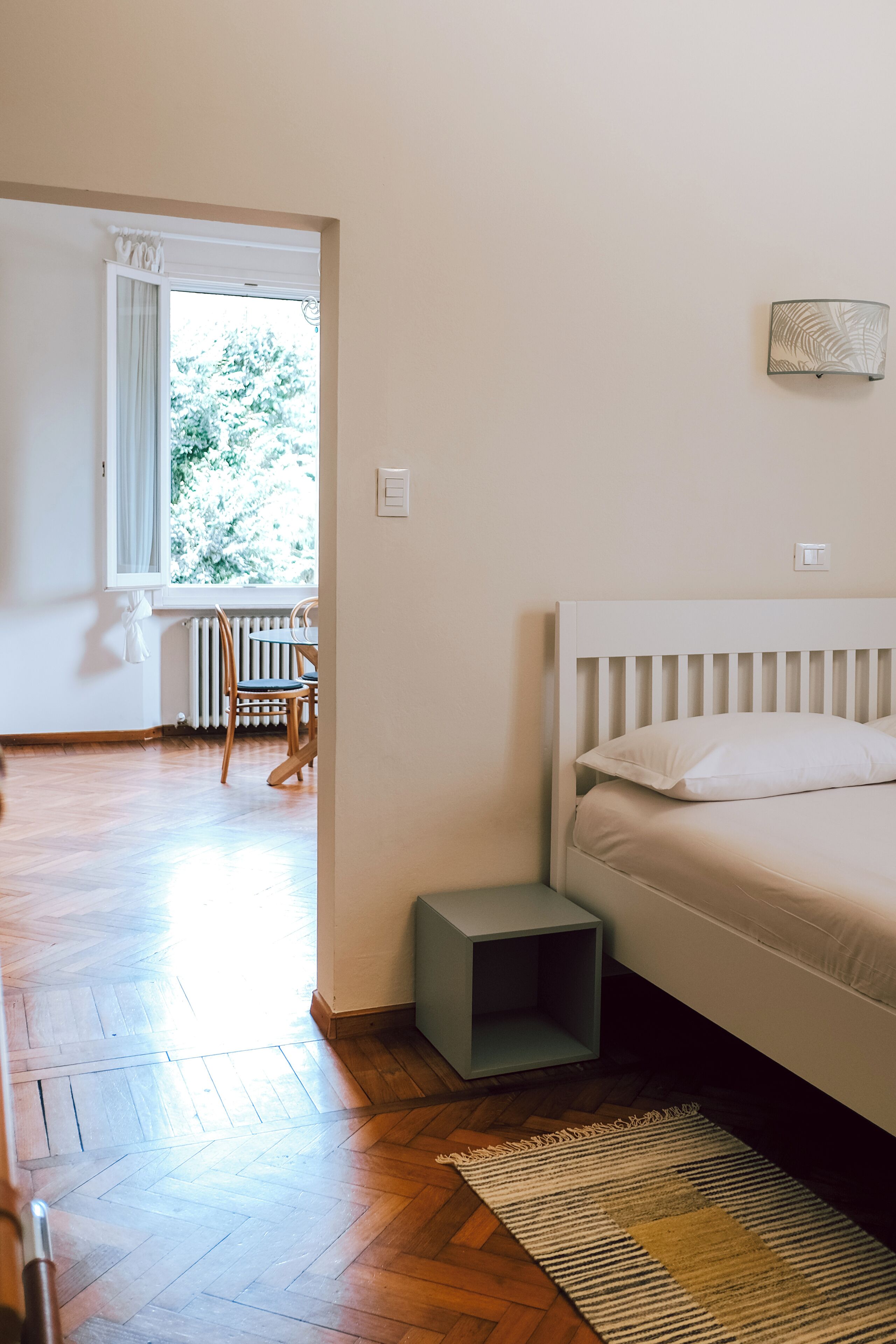 Un dormitorio con una cama, una mesita de noche pequeña, una silla, un radiador y una ventana con cortinas.