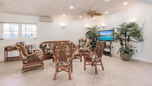 A spacious room with wicker furniture, a ceiling fan, a flat-screen TV, and large windows.
