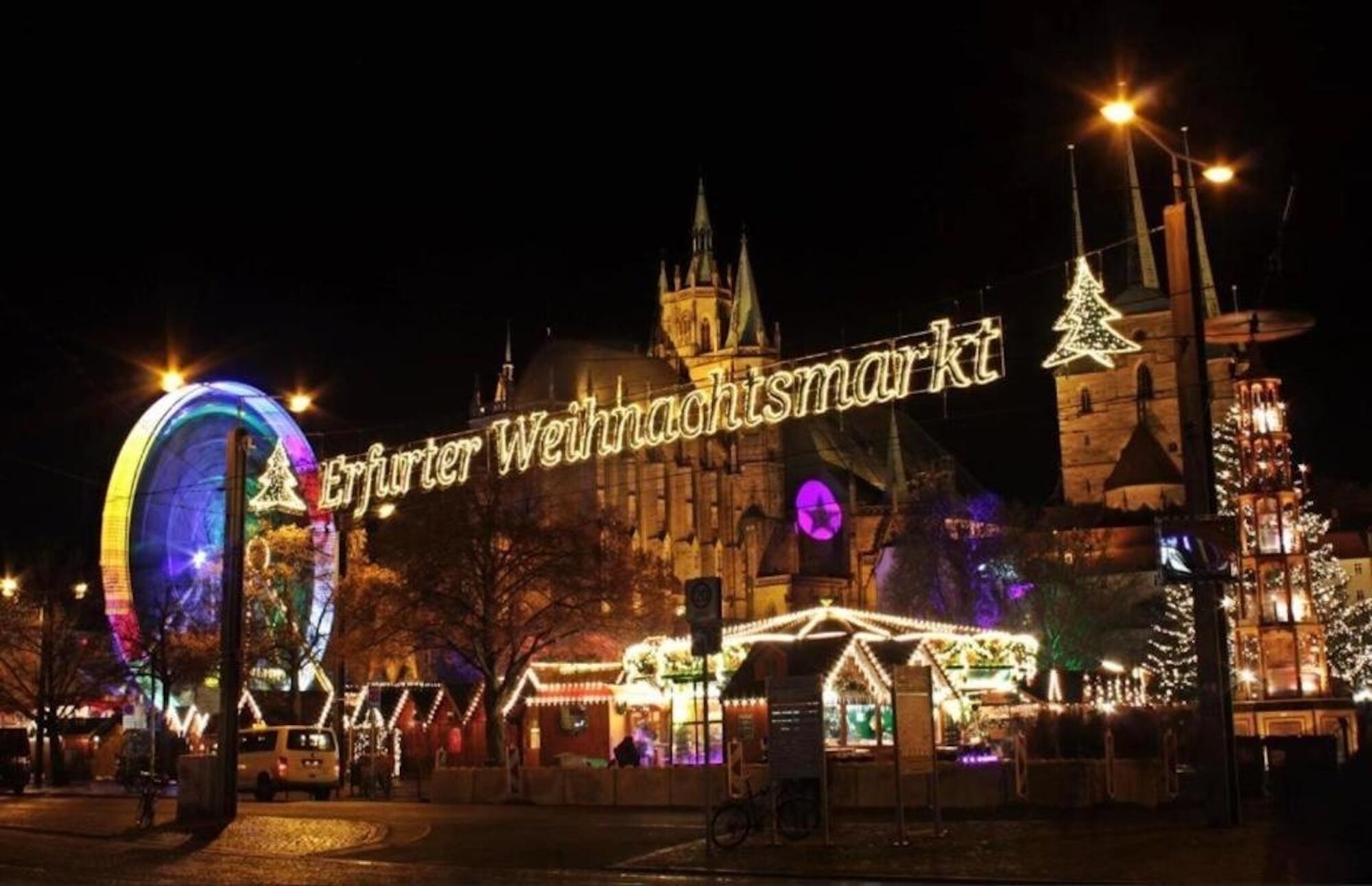 A nighttime view of a historic building with illuminated signage and festive decorations, including a Ferris wheel and Christmas trees.