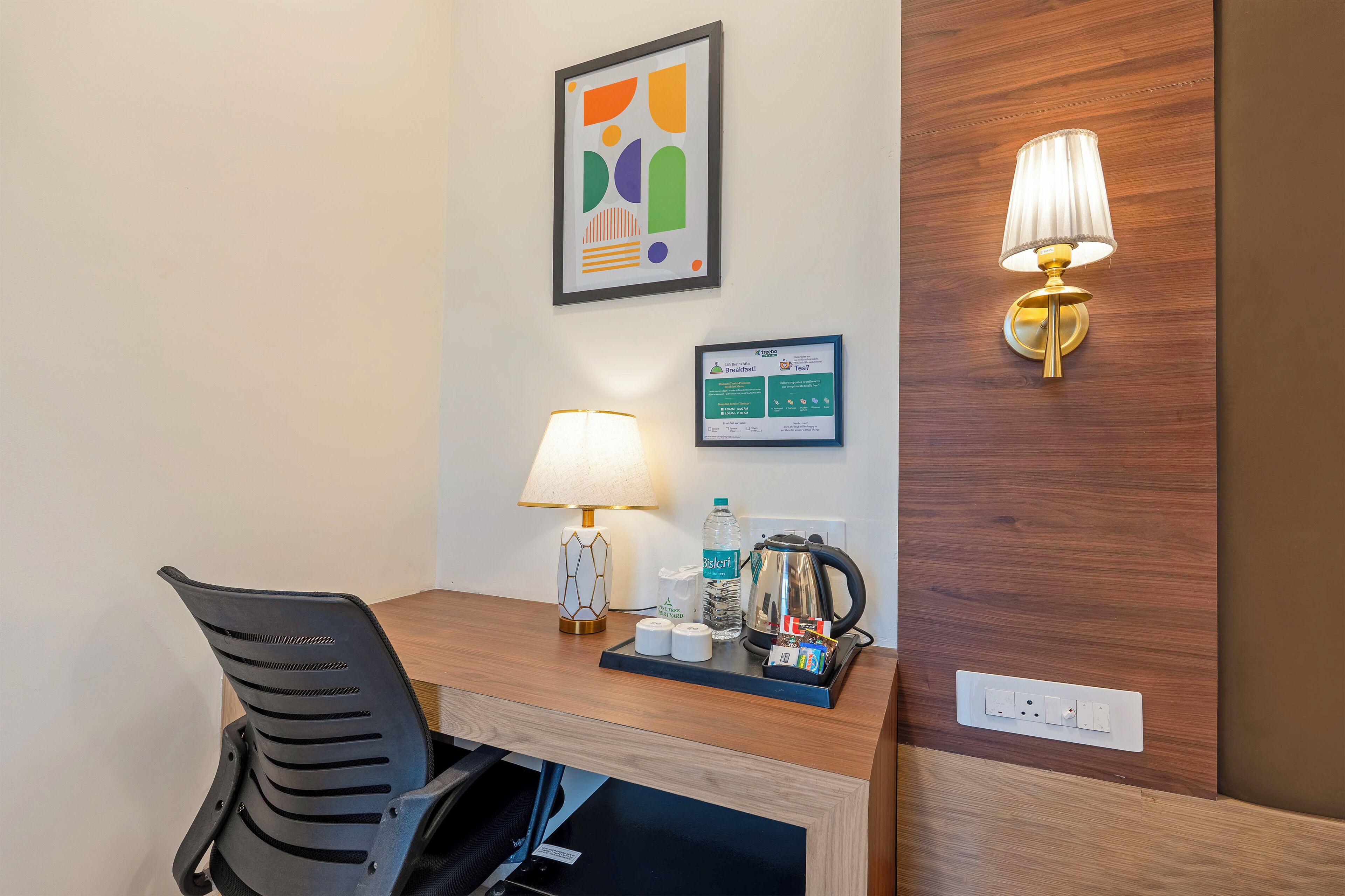 A hotel room with a wooden desk, a black office chair, a lamp, a framed abstract artwork, a bulletin board with a breakfast menu, and a kettle.