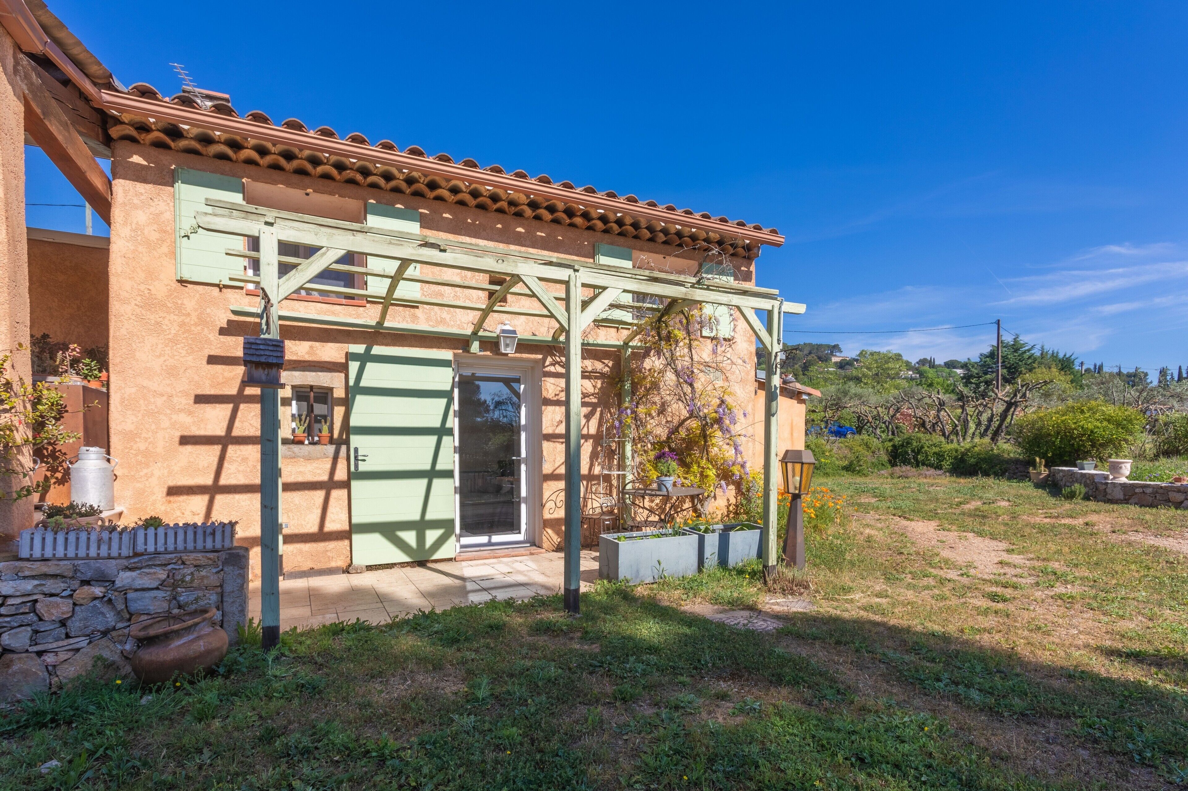 Un edificio rústico con techo de tejas, una pérgola de madera y un muro de piedra con un portón.