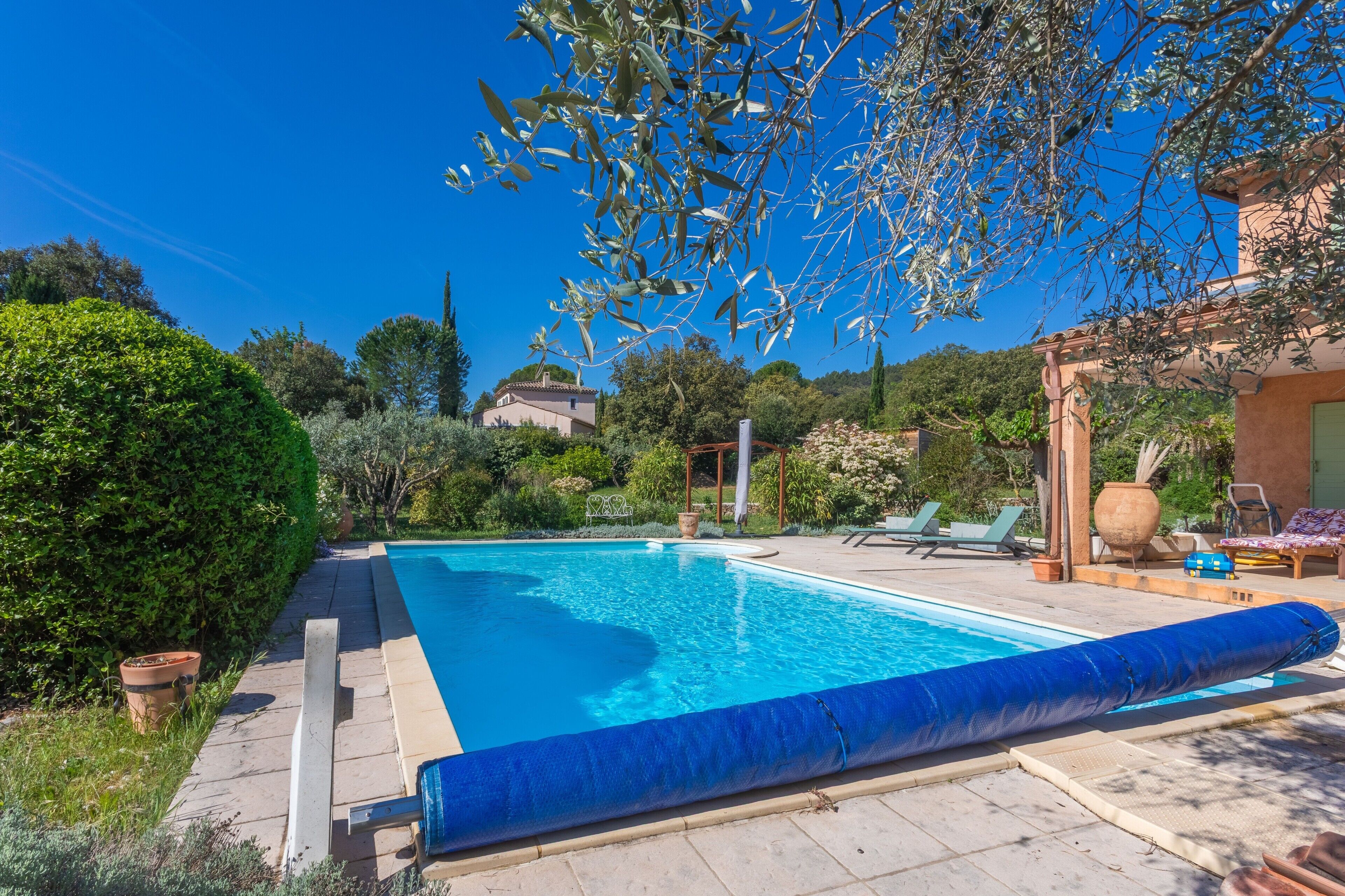 Una piscina con pérgola, sillas de descanso y vistas a una casa al fondo.
