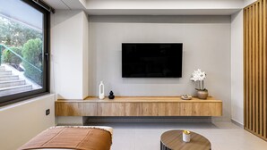 A modern living room with a flat-screen TV mounted on the wall, a wooden bench with decorative items, a large window with a view of outdoor stairs, and a round coffee table. - Clavé - Deluxe Studio in Athens (Glyfada)