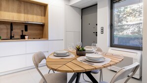A modern kitchen with a wooden table set for dining, white cabinets, and a large window providing a view of the outdoors. - Clavé - Deluxe Studio in Athens (Glyfada)