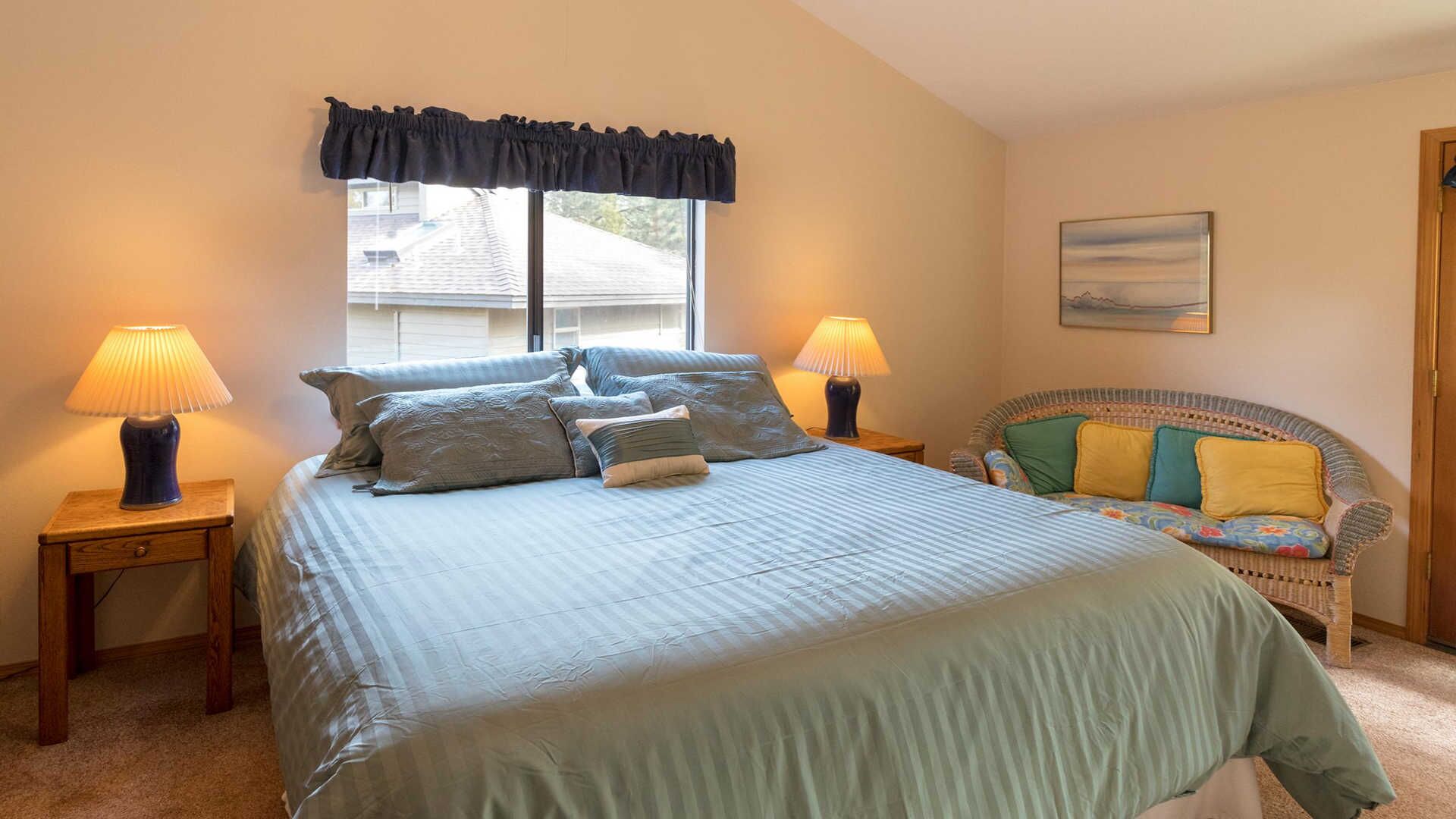 A bedroom with a large bed, two bedside lamps, a window with a valance, a wicker sofa with cushions, and a painting on the wall.