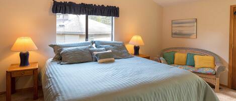 A bedroom with a large bed, two bedside lamps, a window with a valance, a wicker sofa with cushions, and a painting on the wall.