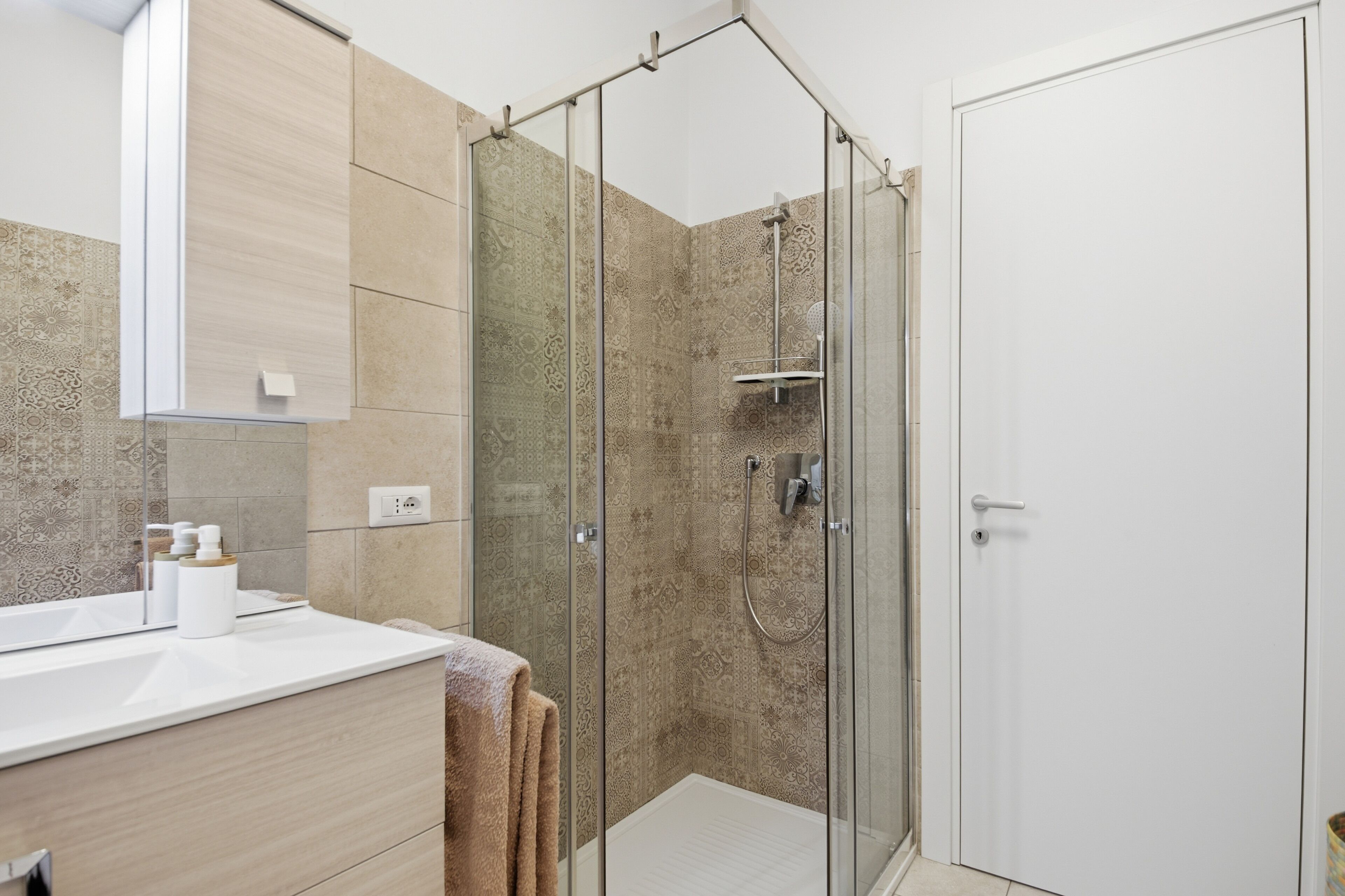 A modern bathroom with a glass shower enclosure, a white sink, and a beige tiled wall.
