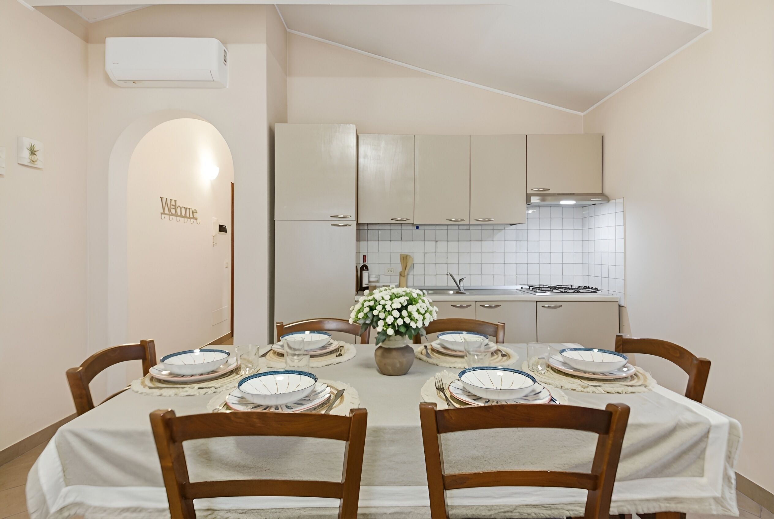 Un comedor con una mesa dispuesta para cuatro personas, rodeada de sillas de madera, un florero con flores y una cocina al fondo.