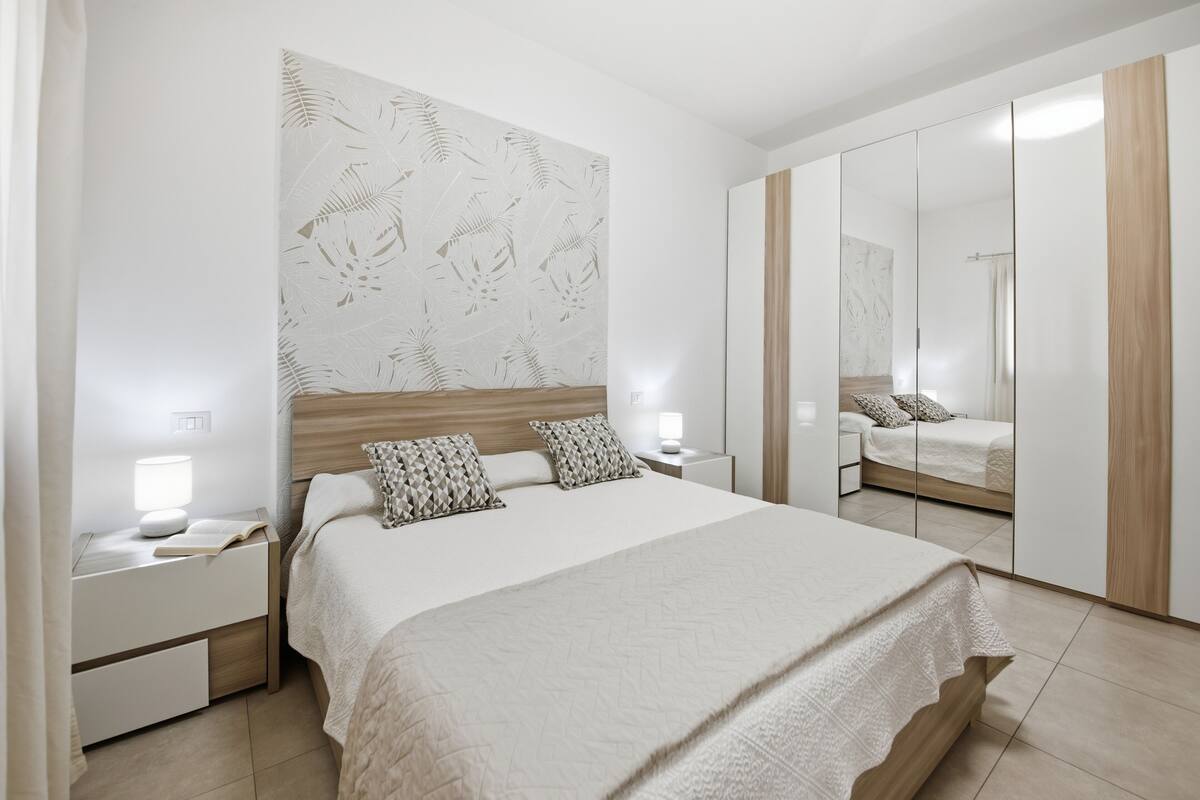 A modern bedroom with a large bed, a nightstand with a lamp, a mirror, and a wardrobe.