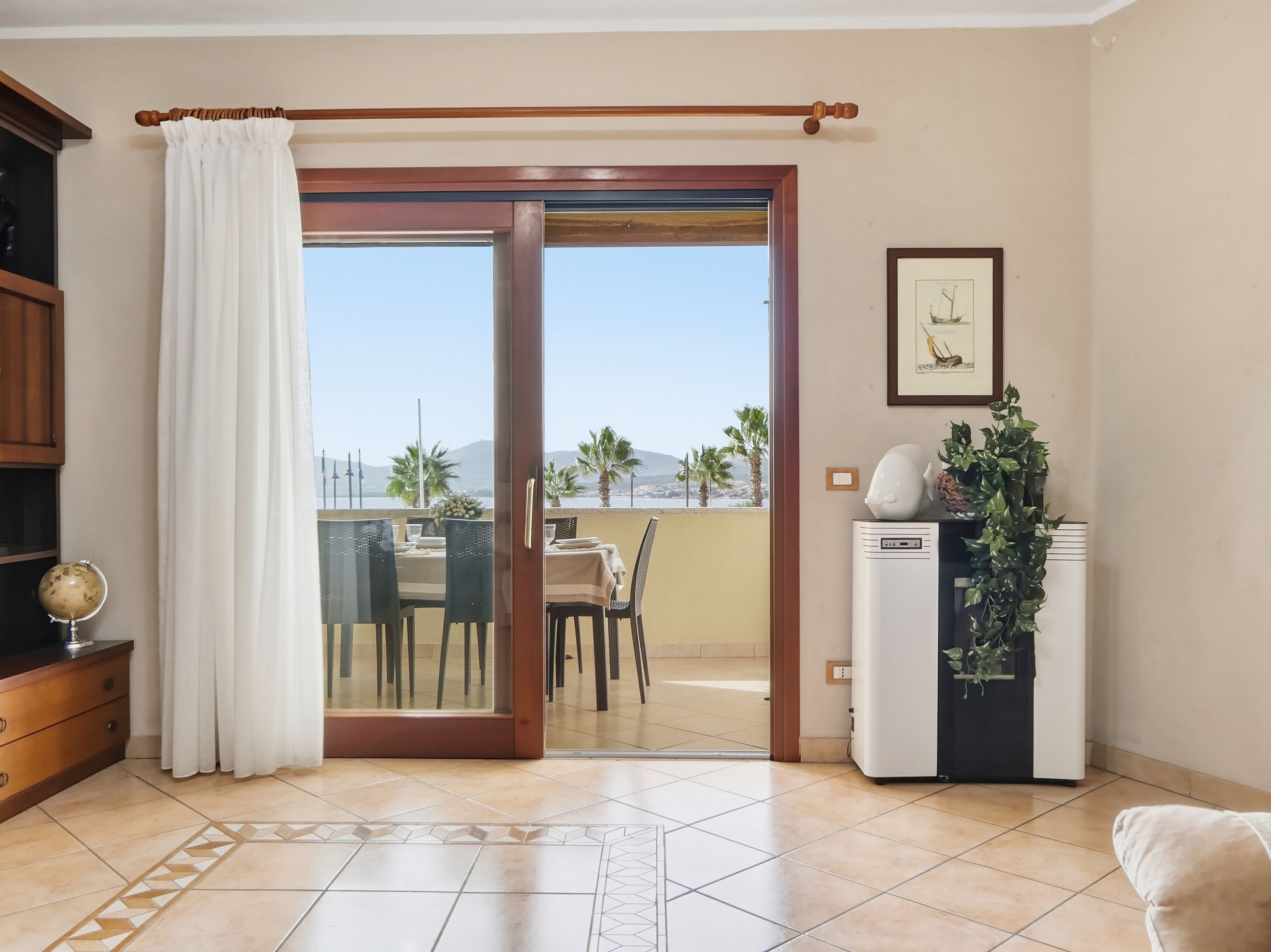 Ein Zimmer mit einer Schiebetür zu einem Balkon mit Blick auf Palmen und Berge. Es gibt einen Esstisch mit Stühlen, ein kleines weißes Gerät und einen Globus auf einer Holzkabine.