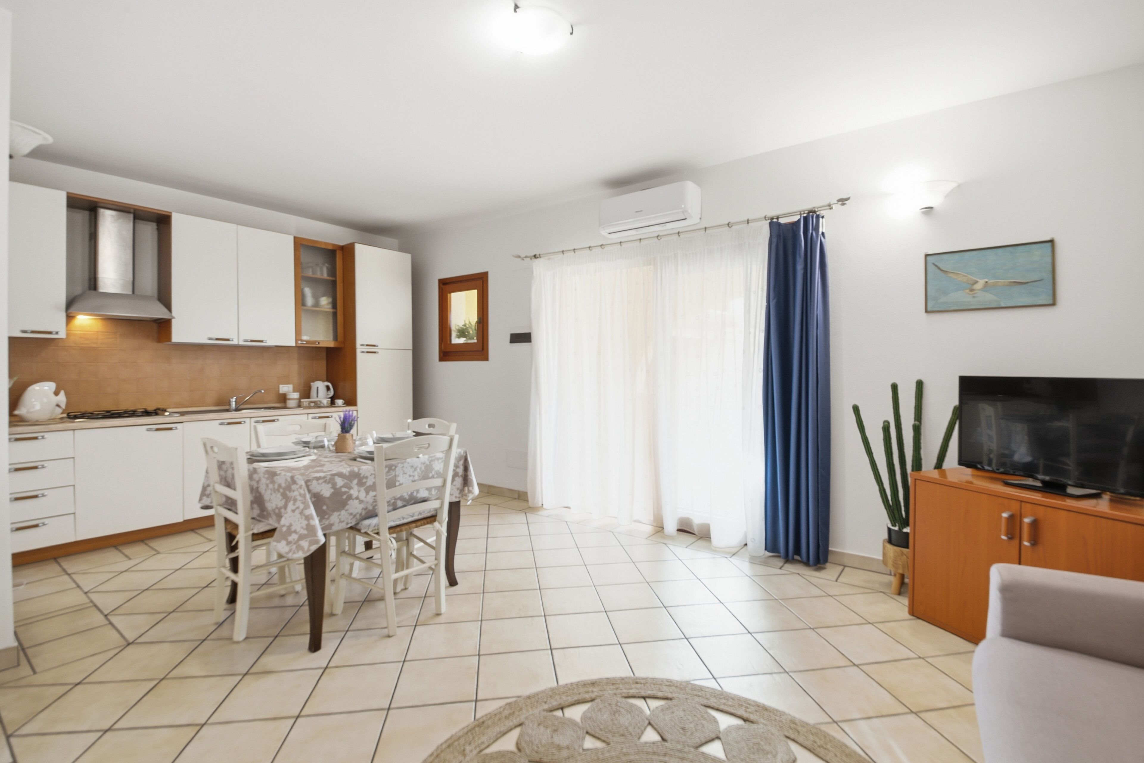 A modern kitchen with white cabinets, a dining table with chairs, a TV on a wooden cabinet, and a large window with blue curtains.