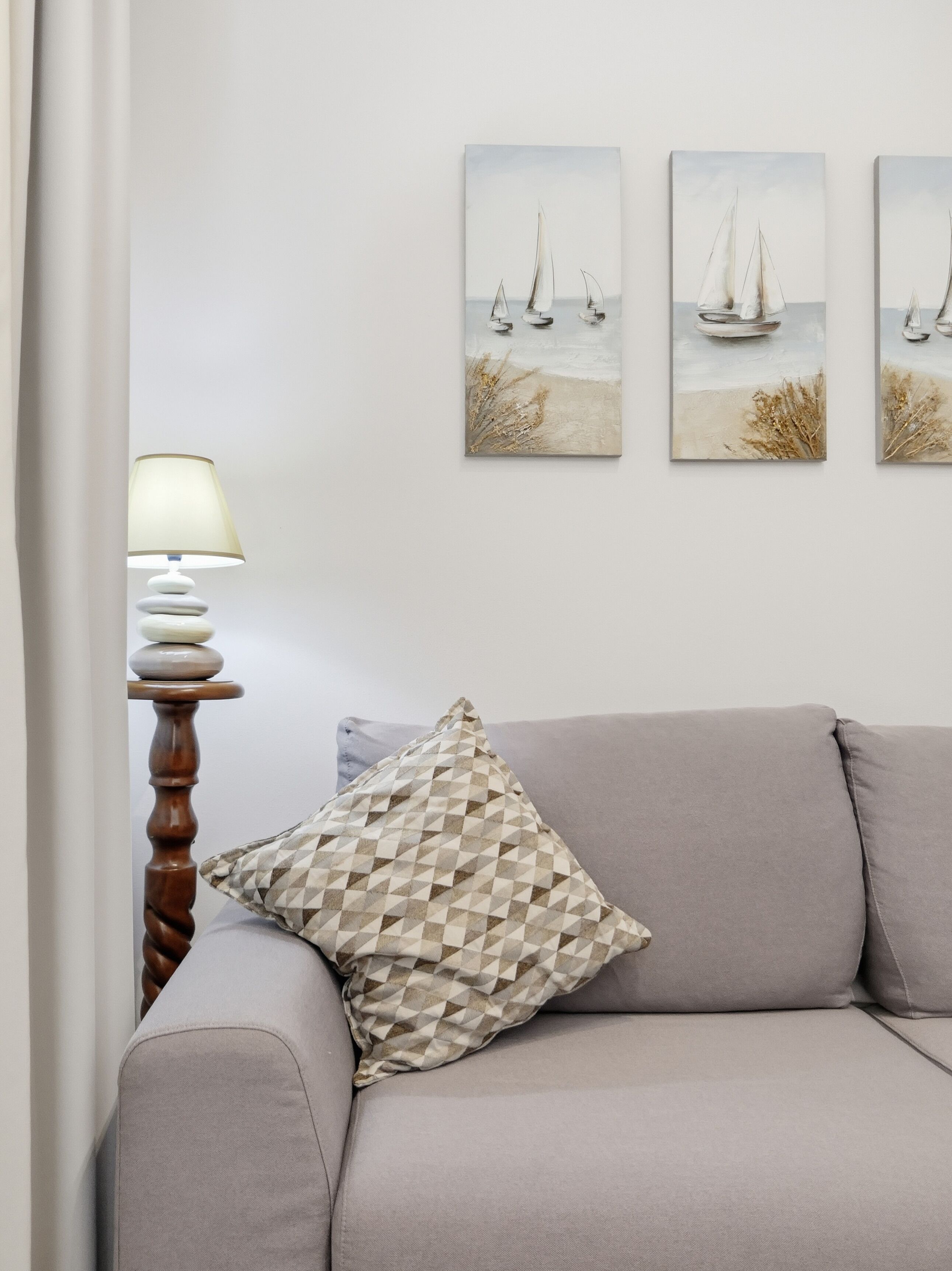 Un soggiorno con un divano grigio, un cuscino decorato, una lampada con libri impilati e tre quadri raffiguranti barche a vela appesi al muro.