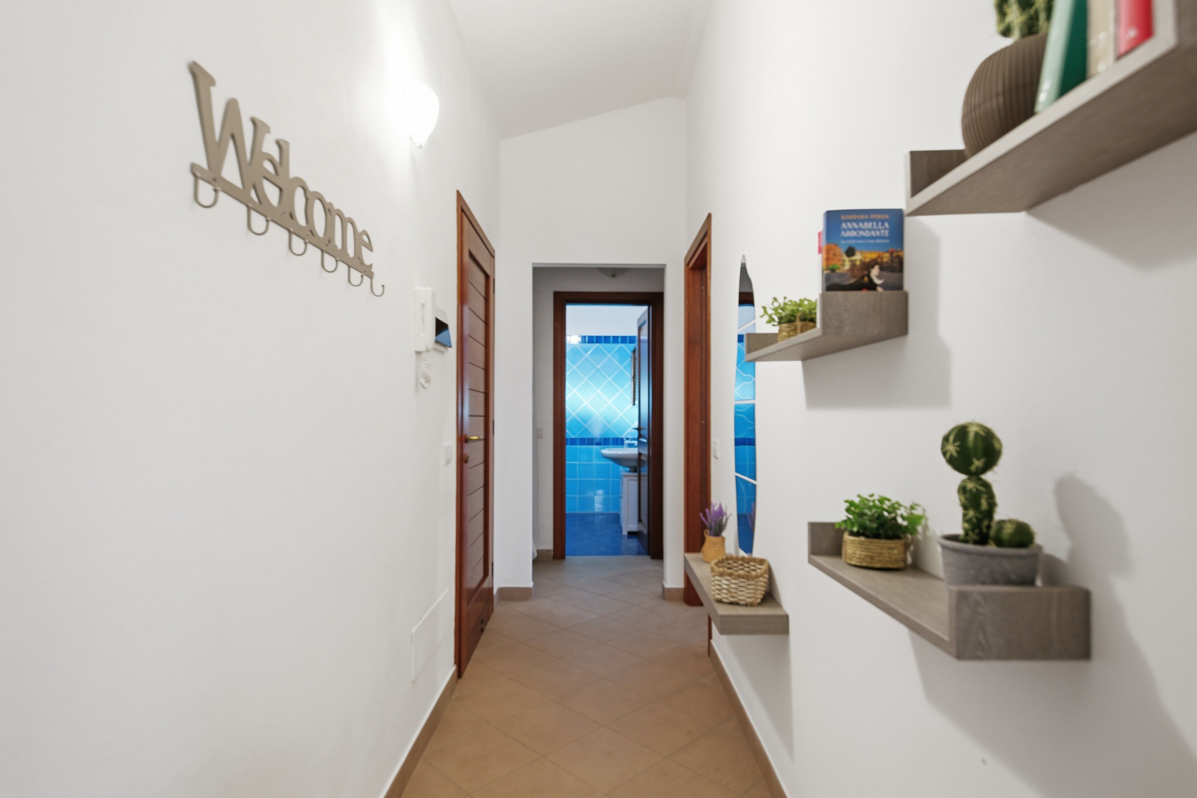 A hallway with a 'Welcome' sign, a door, a shelf with books and plants, and a view into a room with a blue tiled wall.