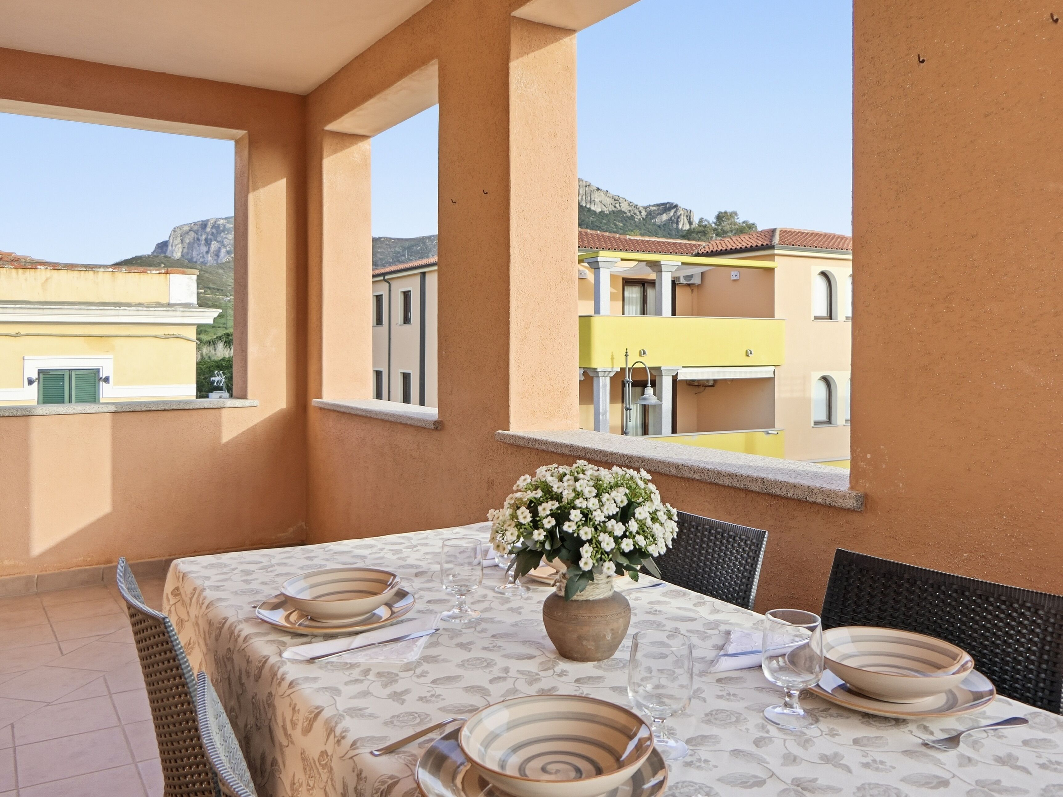 A dining table set with plates, cutlery, and a vase of flowers on a balcony with a view of buildings and mountains.