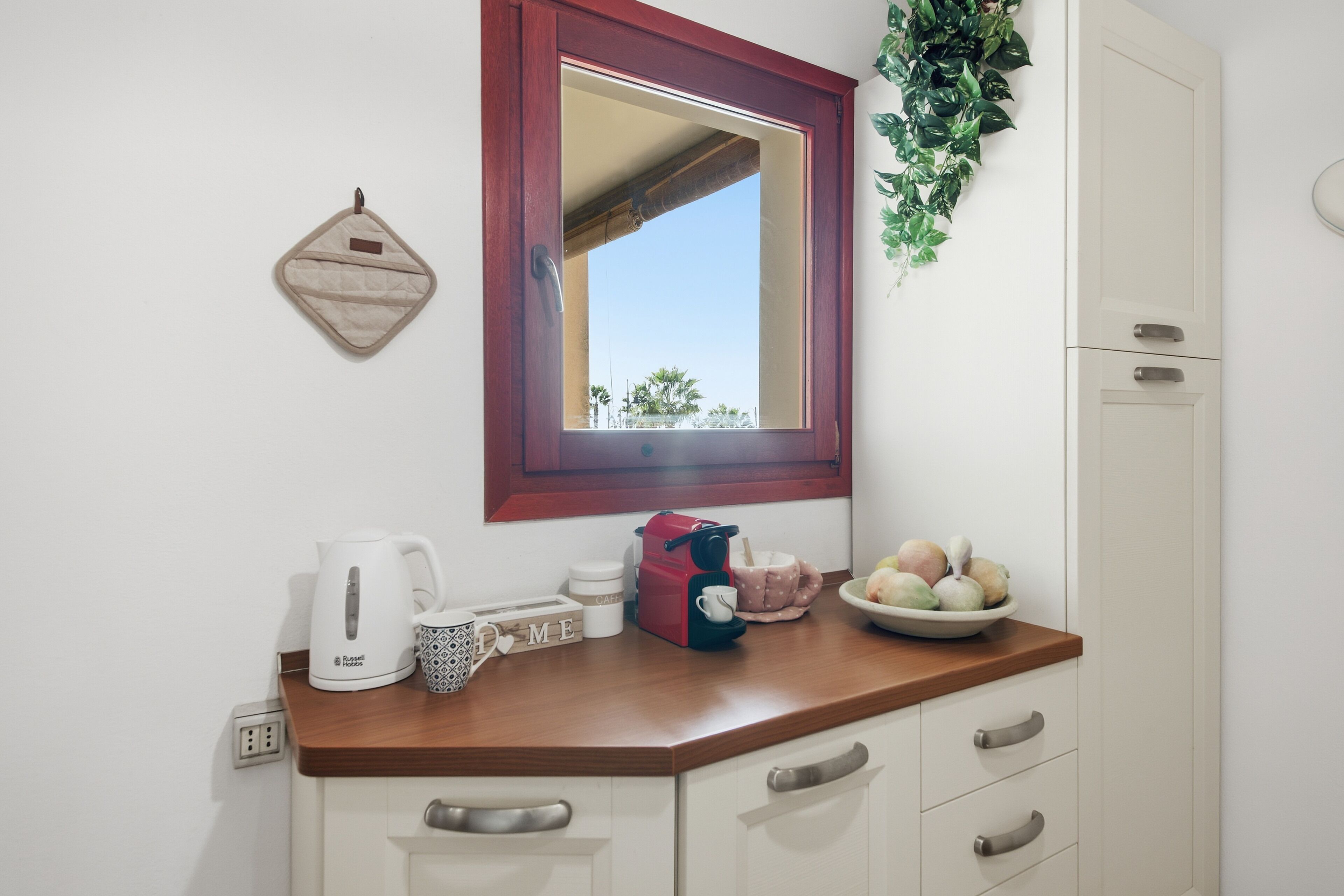 A kitchen with a wooden countertop, a white kettle, a red coffee maker, a bowl of fruit, and a mirror above the counter.