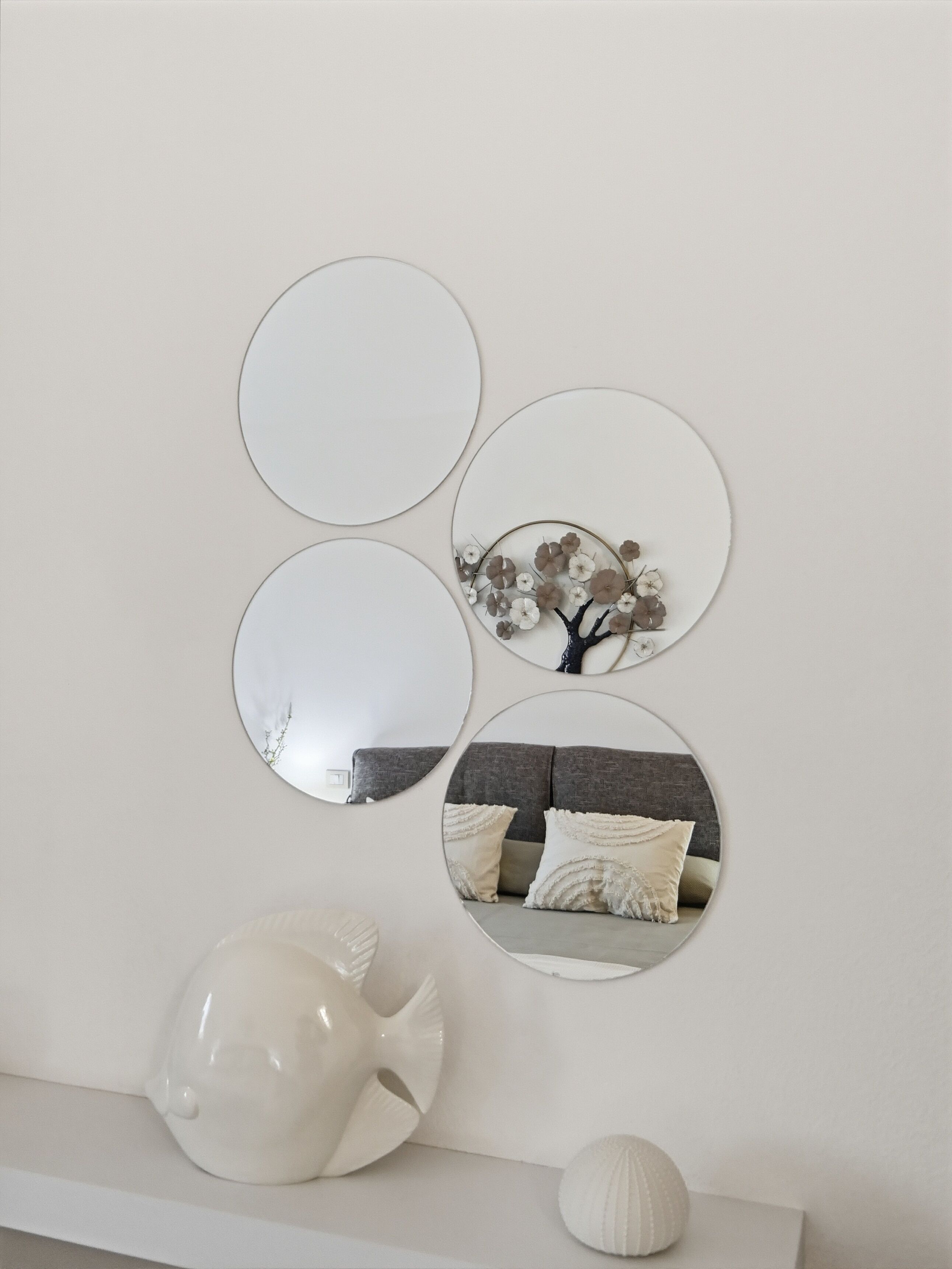 Uma parede com três espelhos circulares de tamanhos diferentes, uma árvore decorativa com flores secas e uma escultura de peixe de cerâmica branca em uma prateleira.