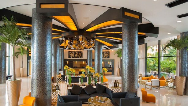 Un hall d’entrée spacieux avec de hauts plafonds, de grandes colonnes et un lustre central. On y trouve des coins salon avec des canapés et une table basse.