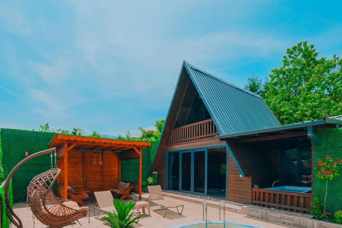 Un espace piscine doté d’une terrasse en bois, d’un fauteuil suspendu et d’un petit bâtiment avec de grandes fenêtres.