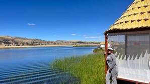 A person standing on a wooden deck overlooking a calm lake with a thatched-roof structure. - Hotel on Lake Titicaca with lake views (Puno)