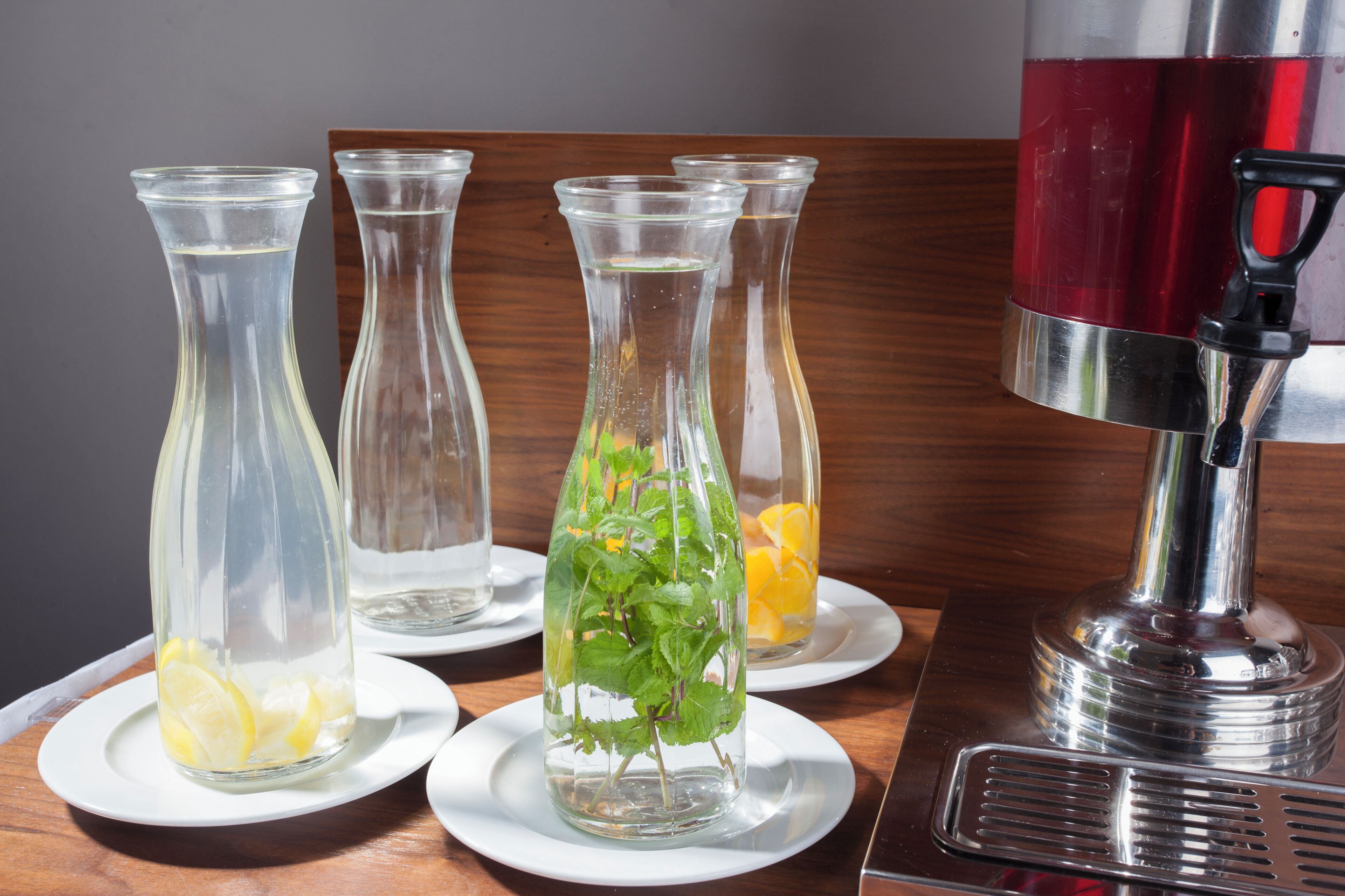Cuatro floreros de vidrio con agua y rodajas de limón sobre platos blancos, y un dispensador de bebidas rojas sobre una mesa de madera.