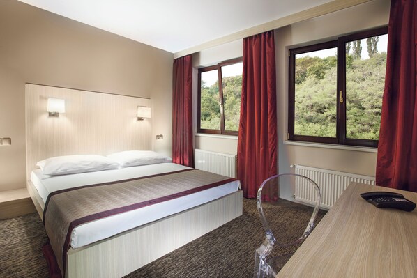 A hotel room with a bed, a desk, a chair, and a window with curtains. - Hotel Hartig (Prague)