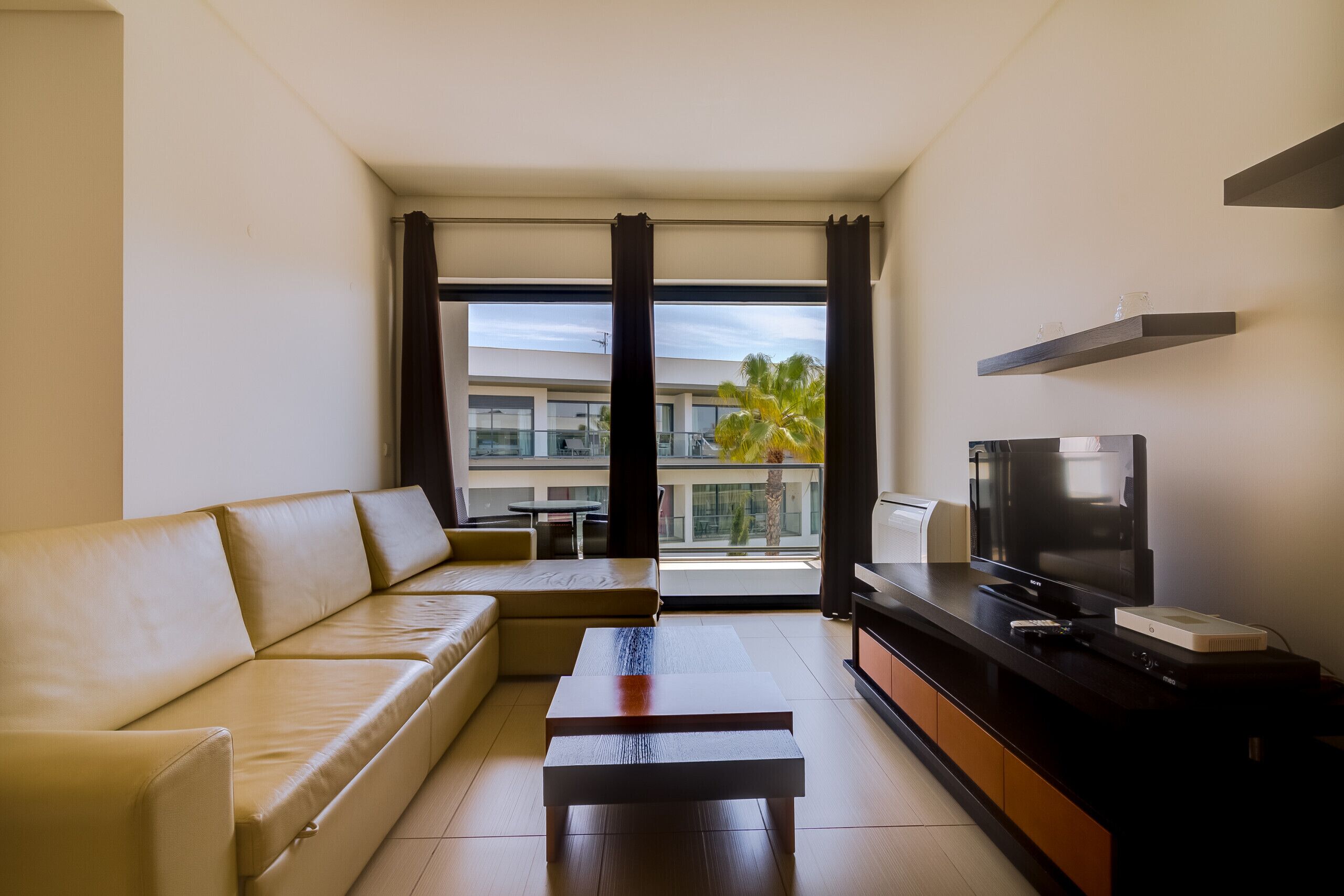 A modern living room with a sofa, coffee table, TV, and a balcony with a view of palm trees.