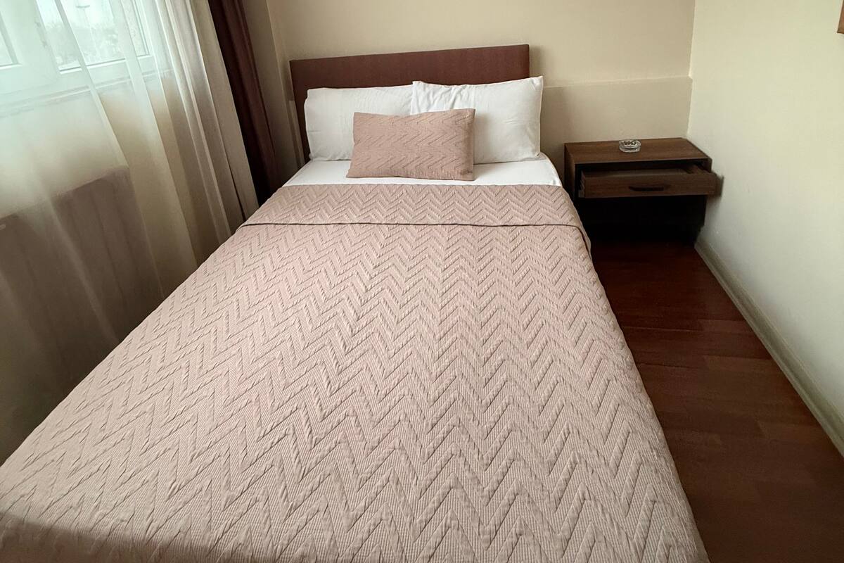 A hotel room with a bed, a nightstand, a window with curtains, and a wooden headboard.