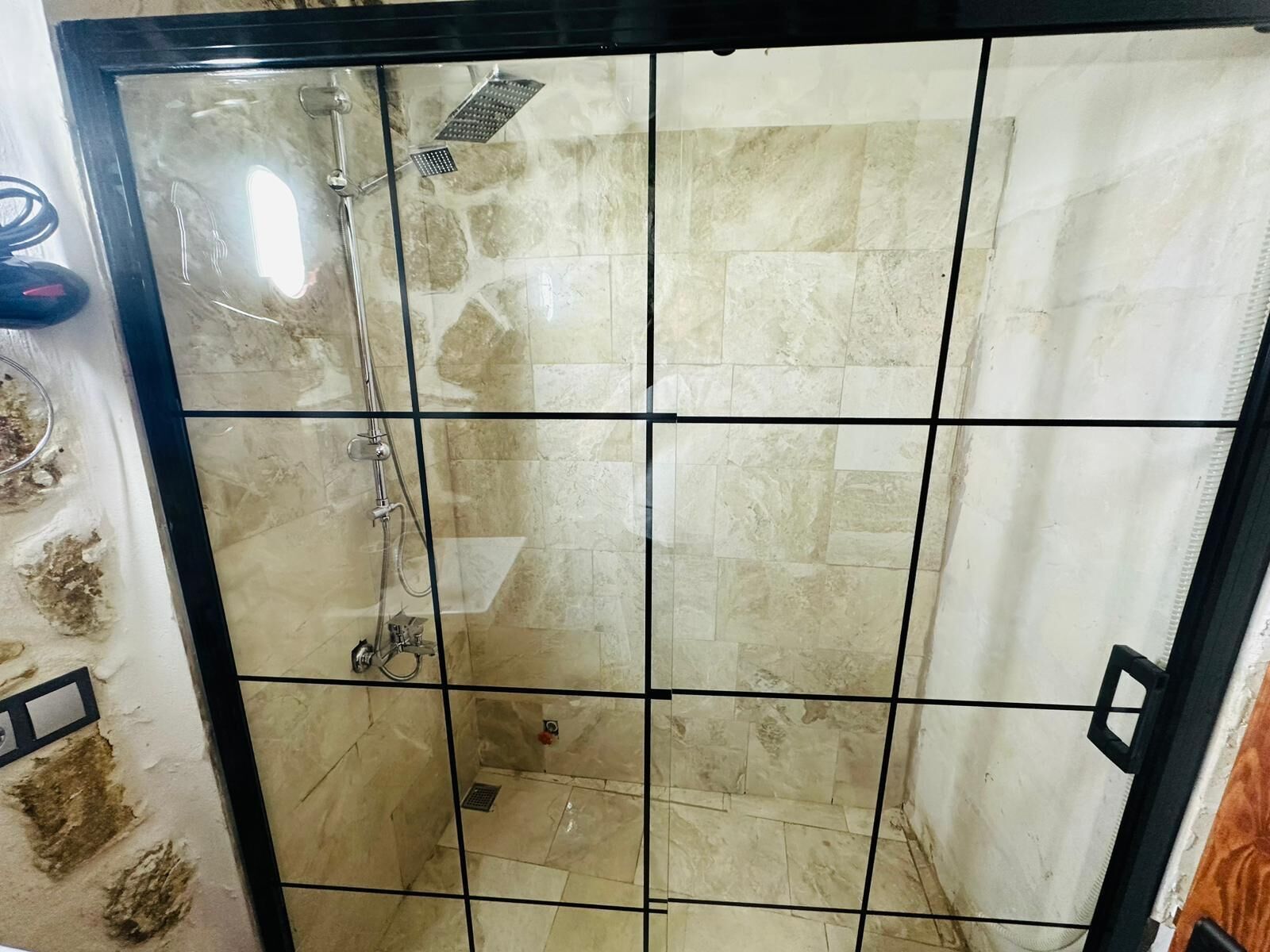 Un área de ducha con paredes y piso de azulejos, mampara de vidrio, cabezal de ducha manual y desagüe.