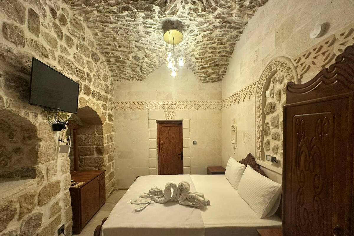 Ein Zimmer mit Steinwänden, einem Bett, einem Holzschrank, einem an der Wand montierten Fernseher und einer Kristallleuchter.