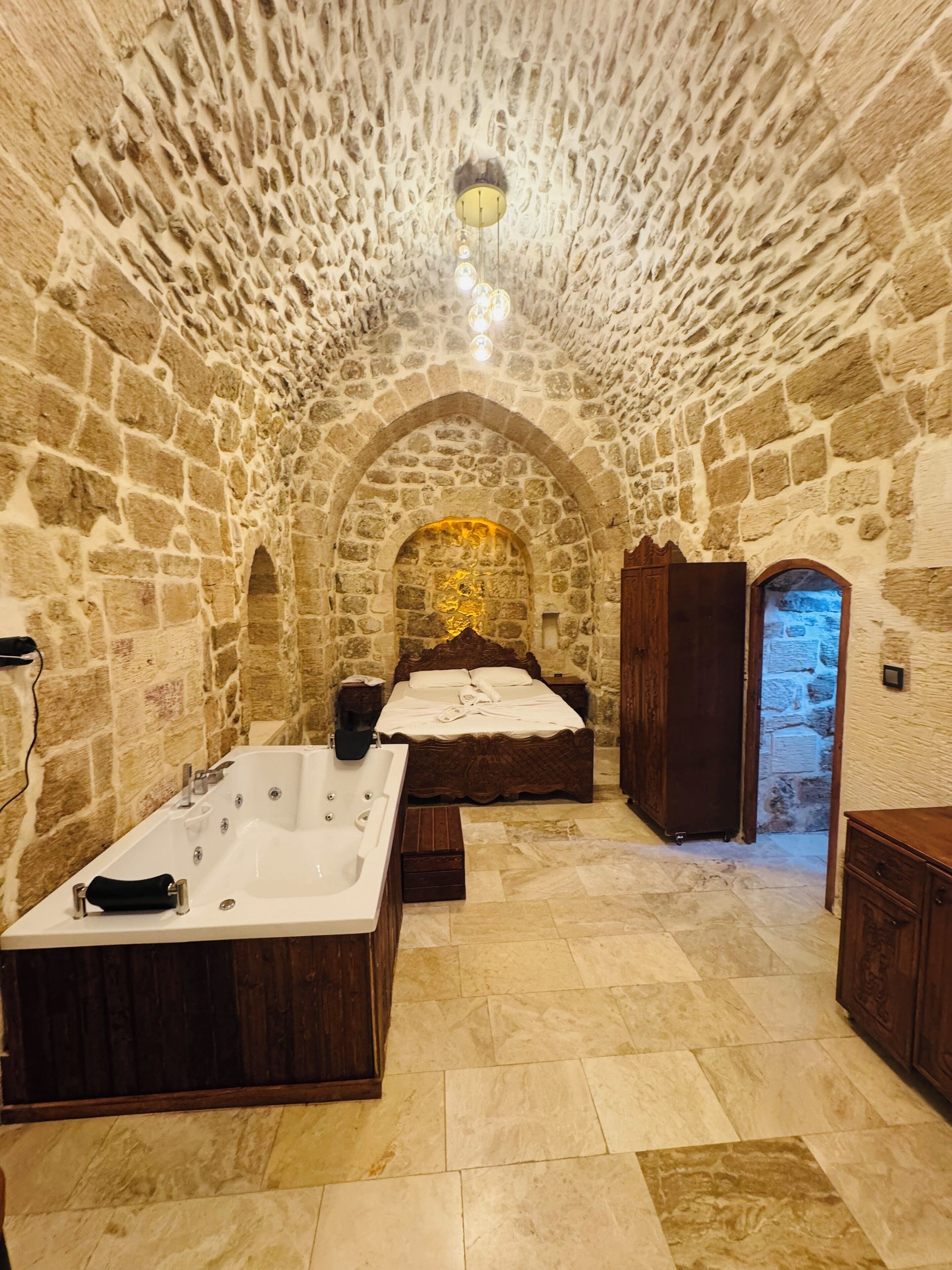 A stone-walled room with a large bathtub, a bed, and a wooden wardrobe.