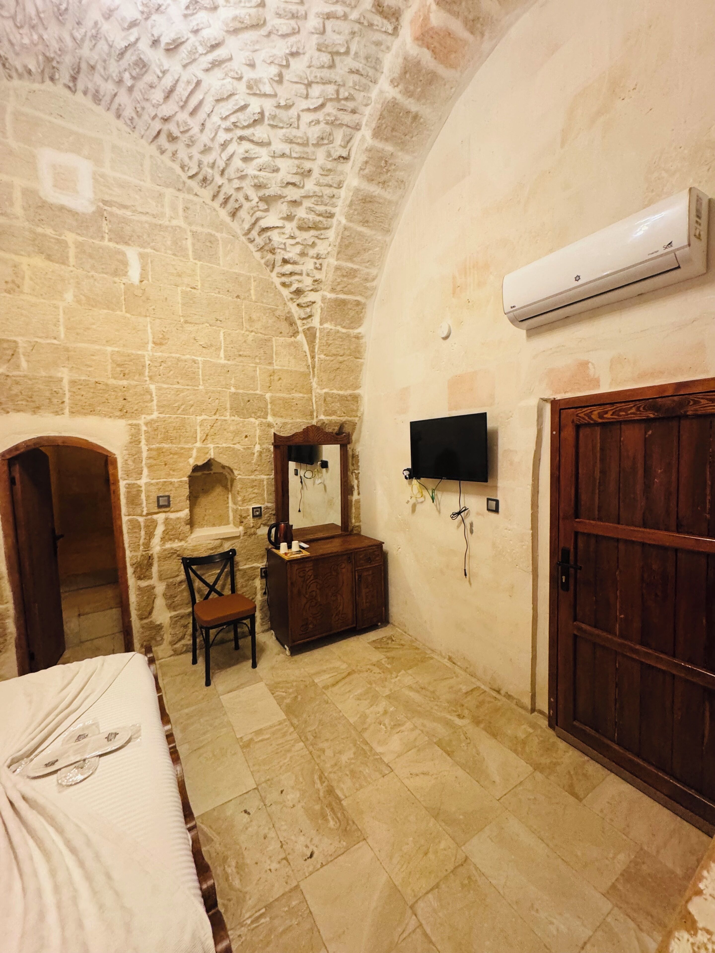 Una habitación con paredes de piedra, una cama, un armario de madera, una silla, un televisor montado en la pared y una puerta de madera.