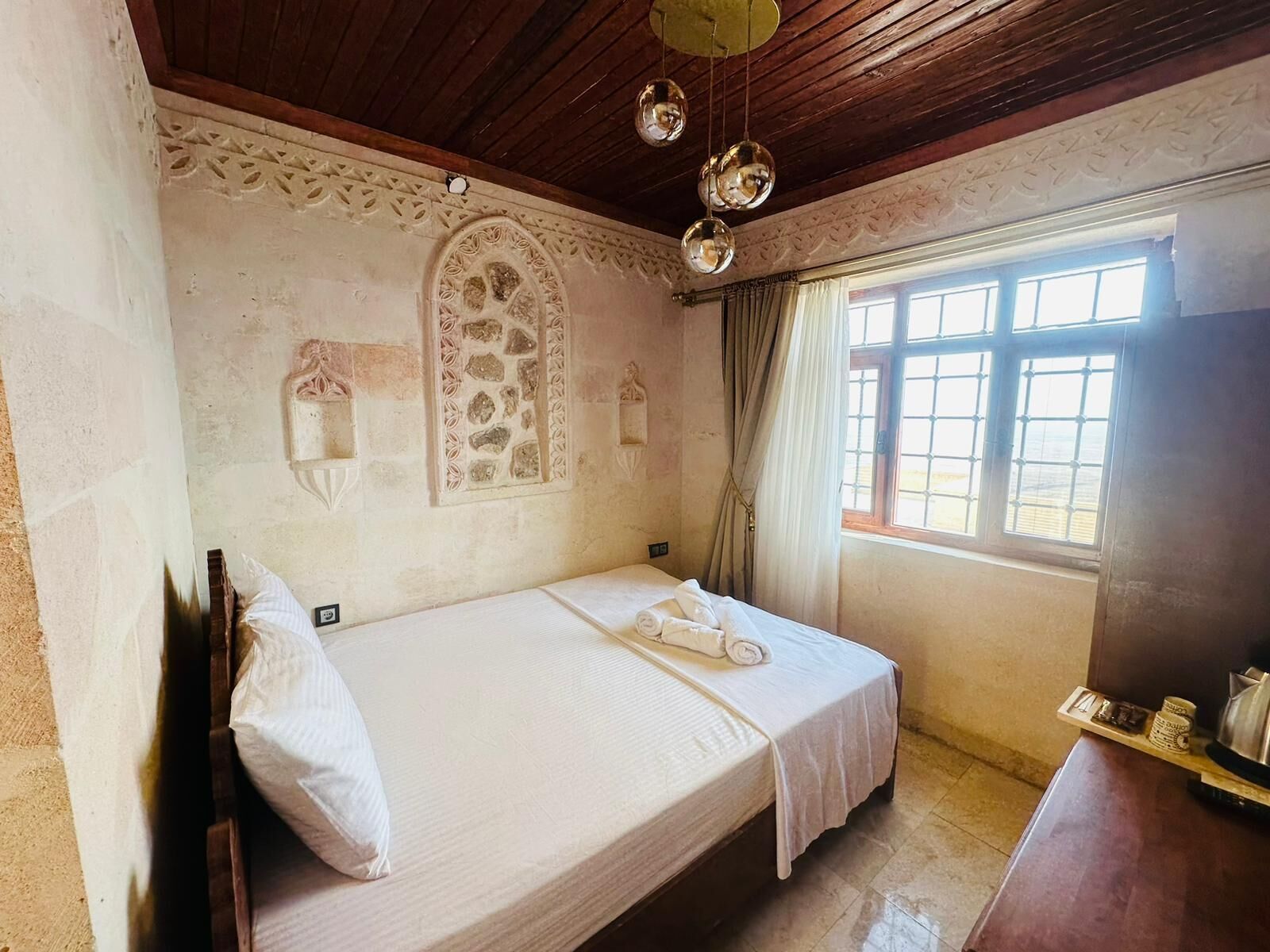 Un lit simple avec un matelas blanc et un oreiller, une tête de lit en bois, une fenêtre avec des rideaux et un plafond en bois orné de luminaires sphériques suspendus.