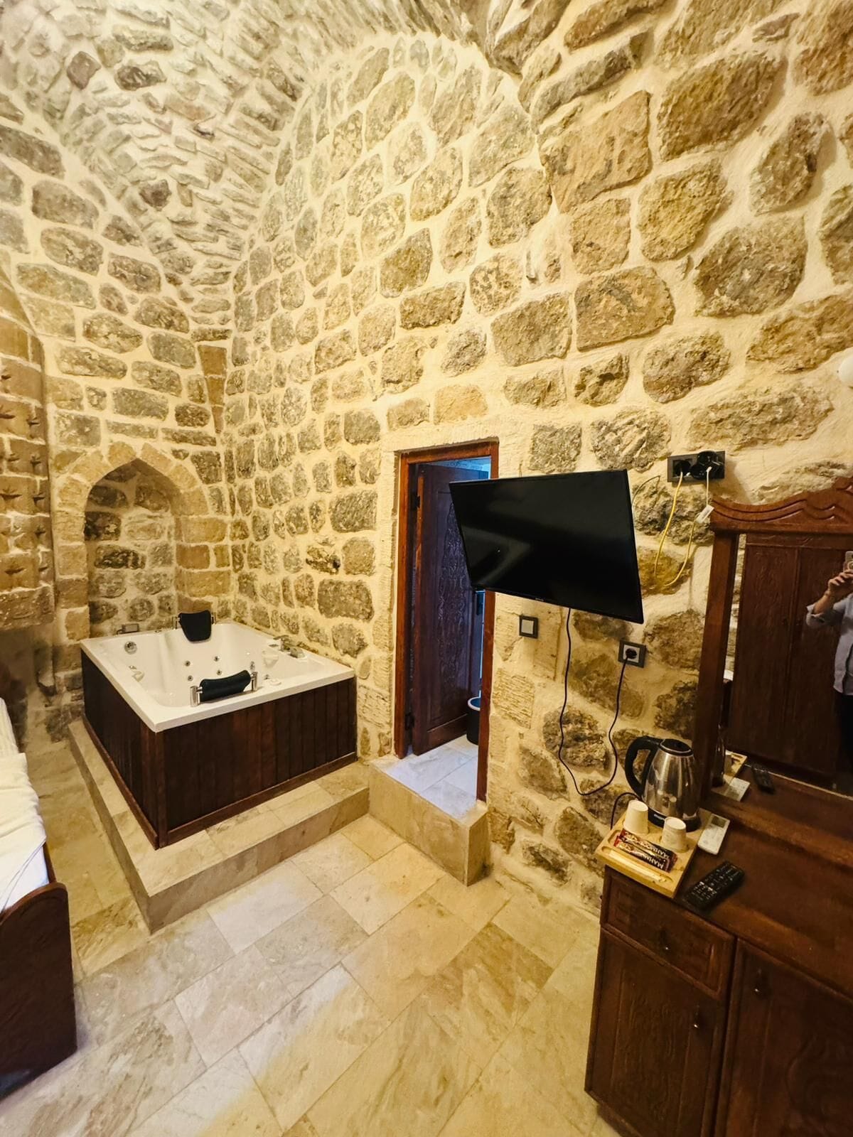 A stone-walled bathroom with a bathtub, a television mounted on the wall, and a wooden cabinet with toiletries.
