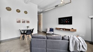 A modern living room with a grey sofa, a dining table with chairs, a flat-screen TV mounted on the wall, and a ceiling with exposed ductwork. - Renovated Downtown Retreat | Pool + In-Unit Laundry (Dallas)