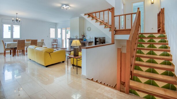 A spacious living area with a yellow sofa, dining table, and chairs, a kitchen area, and a wooden staircase with green and beige tiles. - ALIZEA: House with pool in L'Escala (L'Escala)
