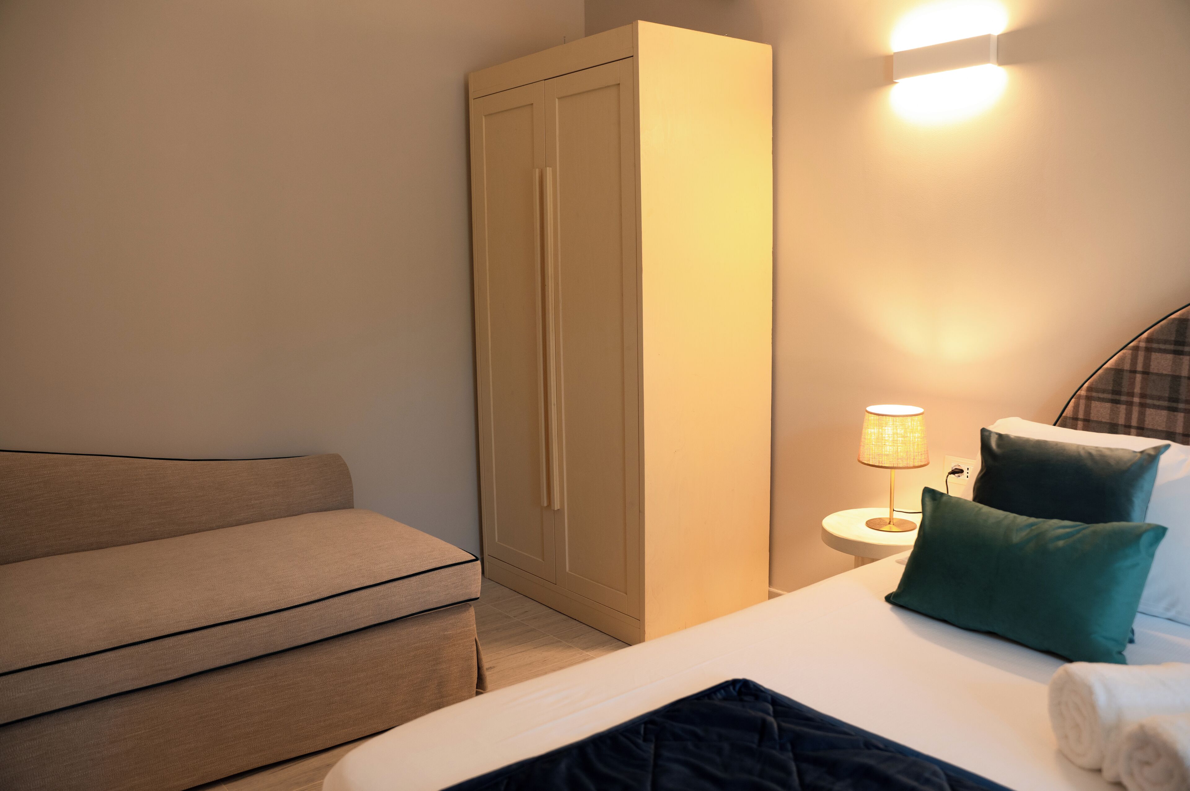 A hotel room with a bed, a wardrobe, a nightstand with a lamp, and a sofa.
