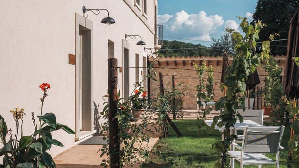 Une cour avec un bâtiment blanc, un chemin en briques et un espace verdoyant agrémenté de chaises et de plantes en pot.