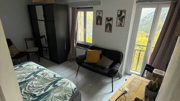 A bedroom with a bed, a sofa, a desk, and a wardrobe. There are windows with curtains and a view of greenery outside.