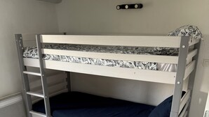 Ein Stockbett mit einer Leiter, einer gemusterten Matratze und einem grauen Teppich in einem Raum mit weißen Wänden und einer Holzbalkendecke.