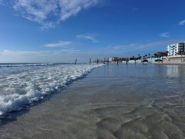 Ein Strand mit Wellen, blauem Himmel und Gebäuden im Hintergrund.