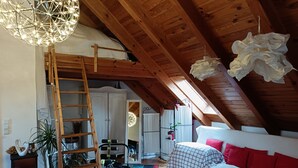 A loft bedroom with a wooden ceiling, a colorful rug, a white sofa with red pillows, and a ladder. - Apartment "Blütenlicht" with Mountain View, Shared Terrace and Wi-Fi (Hameln)