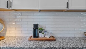 Una cocina con armarios blancos, encimera de granito, cafetera, planta en maceta y tabla de cortar de madera.