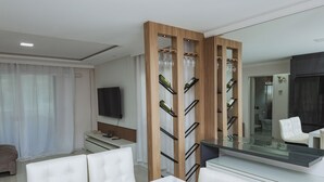 A modern living room with a white sofa, a glass-top coffee table, and a wooden wine rack with bottles and glasses. - House with pool table and barbecue - 10 min from Balneário Camboriú! (Camboriú)