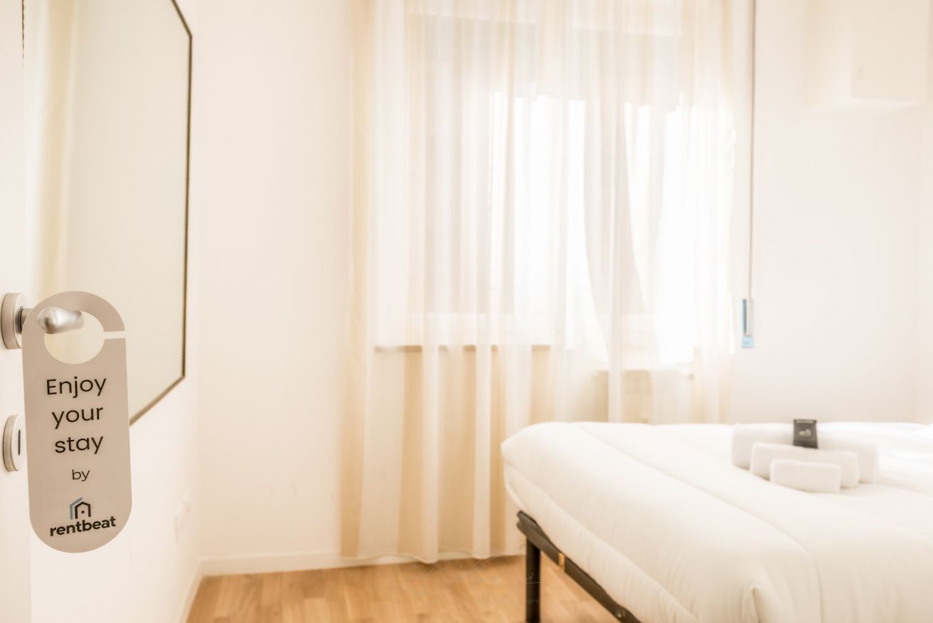 A hotel room with a bed, a window with sheer curtains, a door with a sign, and a wooden floor.