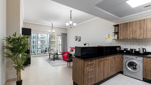 A modern kitchen with wooden cabinets, a built-in washing machine, and a dining area with a red sofa. - Maison Privee - Modern Downtown Apartment w/ City Skyline Vws (Dubai)