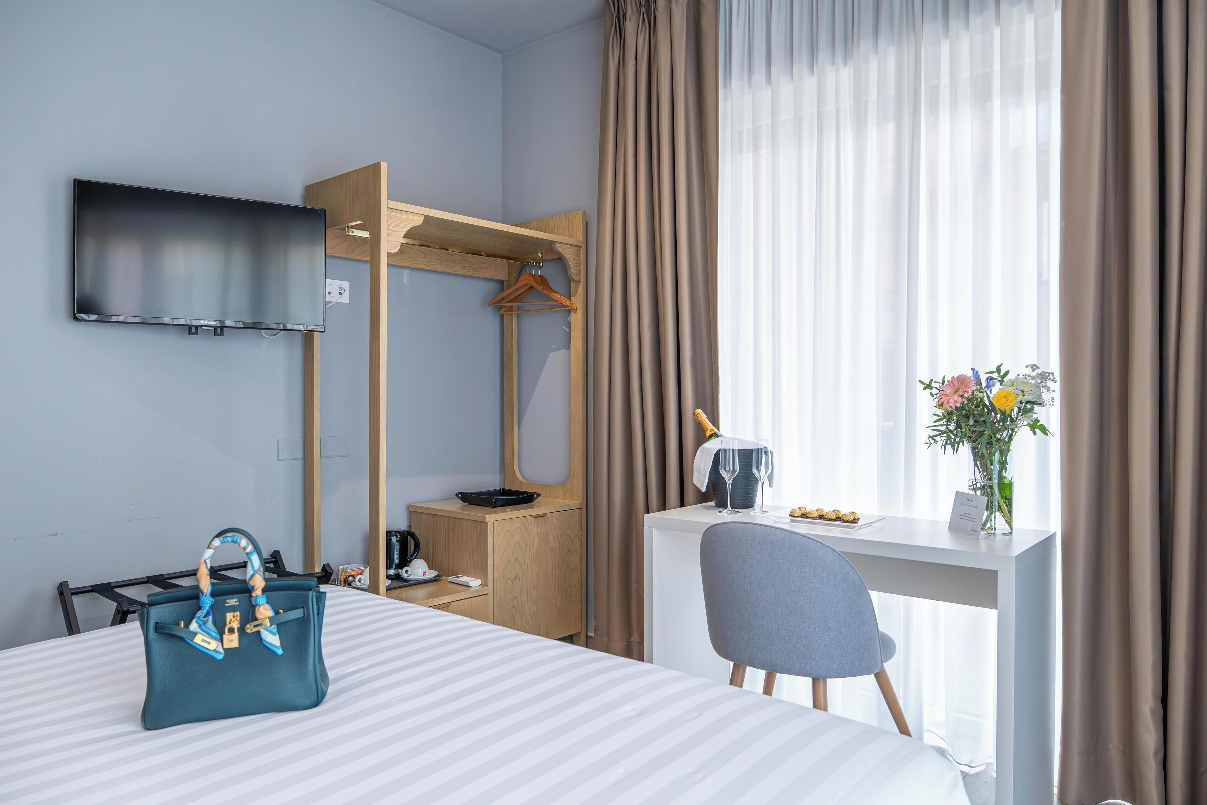 Een hotelkamer met een bed, een bureau met stoel, een aan de muur bevestigde televisie, een houten garderobe en een raam met gordijnen.