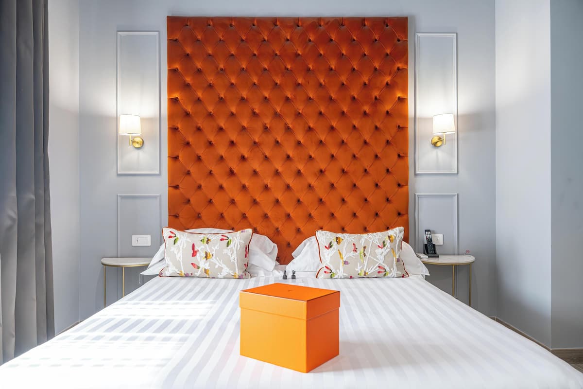 Habitación de hotel con un cabecero capitonado, dos mesitas de noche con lámparas, una cama con ropa de cama a rayas y una pequeña caja naranja sobre la cama.